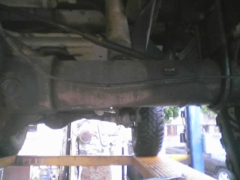 1990 Ford Bronco - Undercarriage view of a rear differential and driveshaft with a mud tire and shop lift post visible, taken under a raised vehicle.