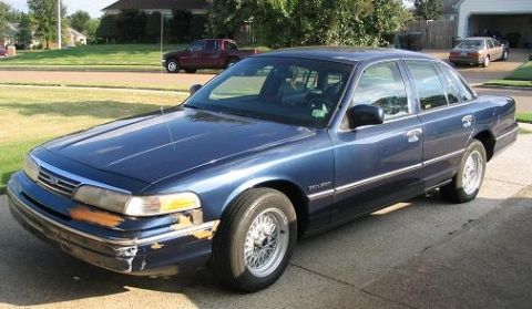1994 Ford Crown Victoria - A blue 1994 Ford Crown Victoria sedan is parked on a driveway, with a red pickup truck and another car visible in the background.