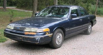 1994 Ford Crown Victoria - A 1994 Ford Crown Victoria is shown from the front three-quarter view on a gravel surface with trees in the background.