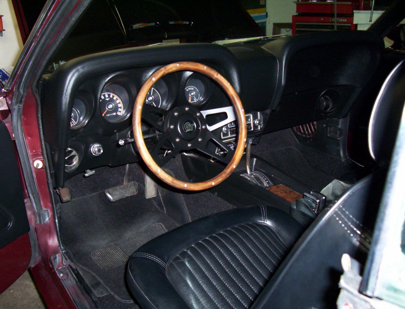1969 Ford Mustang interior with wood GT steering wheel, black gauges and black vinyl seats, seen through open driver door