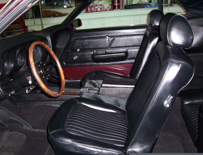 Black interior of a 1969 Ford Mustang with wood steering wheel and vinyl bucket seats, seen through open door