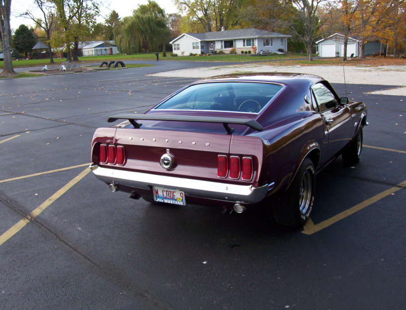 Maroon 1969 Ford Mustang with a rear spoiler and chrome bumper, viewed from the rear quarter