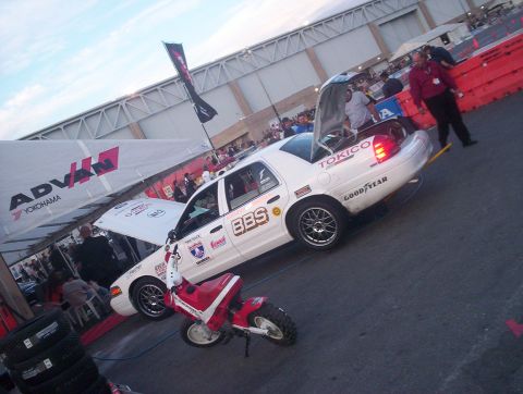 1998 Ford Crown Victoria - white Ford Crown Victoria covered in sponsor decals including BBS, Tokico and Goodyear, parked with a red mini bike at an outdoor event.