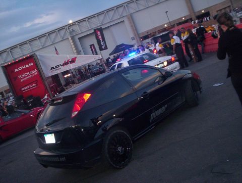 1998 Ford Crown Victoria - black Ford Focus hatchback with PowerHouse decals and black wheels parked at a dusk motorsport event with a police car behind.
