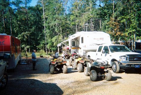 2003 Ford F-350 - A white Ford F-350 truck is parked next to a camper, with ATVs and a trailer in a wooded area.