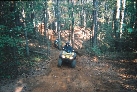 2003 Ford F-350 - Two ATVs navigate a muddy trail through a dense forest, with one rider in the foreground and another further up the path.