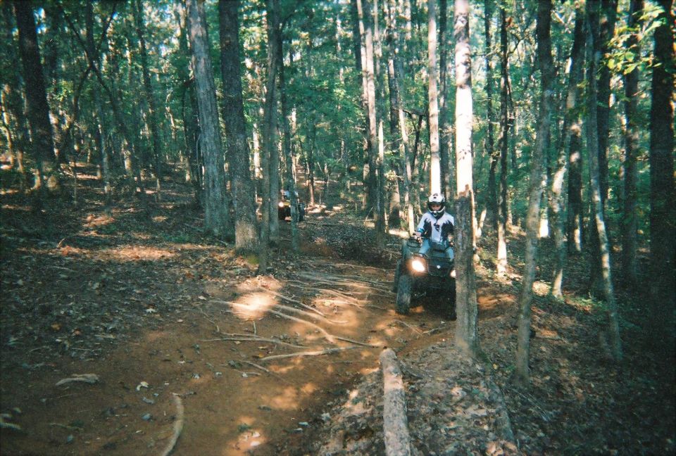 Rider on an ATV on a dirt trail in the woods. Another ATV is visible further back on the trail