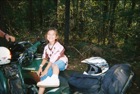 2003 Ford F-350 - A young girl smiles while sitting on a green Kawasaki ATV, surrounded by trees and camping gear.