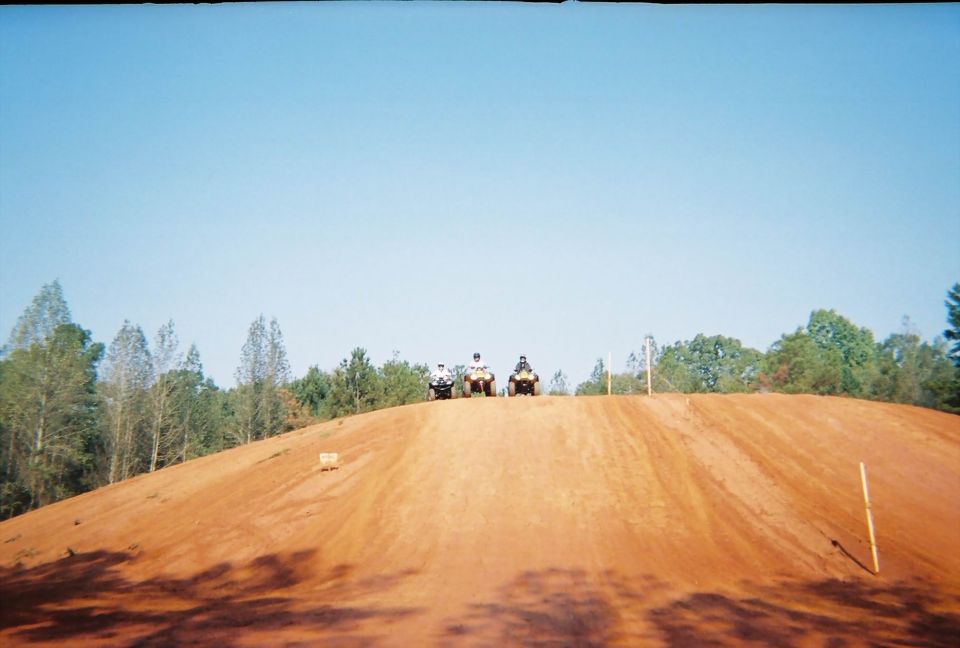 Three ATVs are lined up on top of a dirt hill, with trees in the background and a clear blue sky