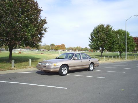 1993 Ford Crown Victoria - A tan sedan is parked in an empty parking lot next to a grassy field with trees.