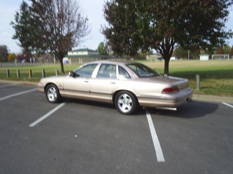 1993 Ford Crown Victoria - Side view of a tan sedan parked in a lot with trees and a building in the background.