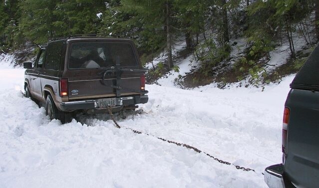 Brown two-door SUV stuck in snow, chained to a teal SUV for towing in a forest.