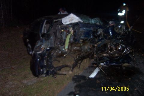 1998 Ford Crown Victoria - Night scene of a dark sedan destroyed in a severe collision on a roadside, with the entire front clip torn away, engine and suspension comp…