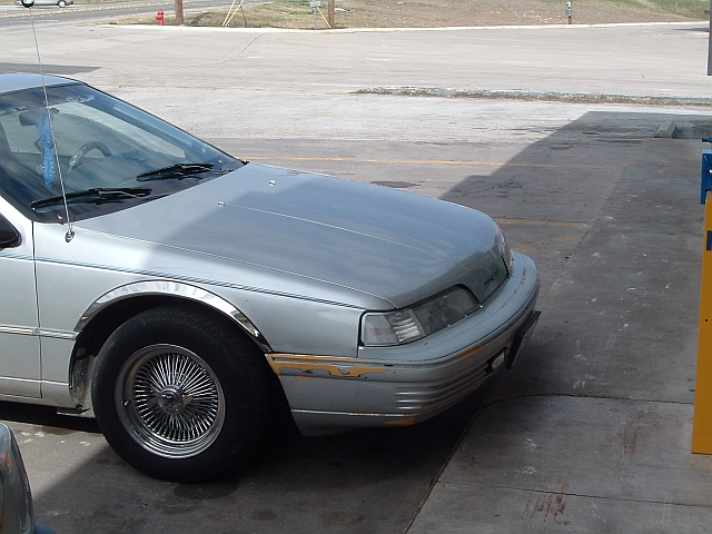 Silver sedan with wire wheels parked on concrete. Front fender and bumper show some damage