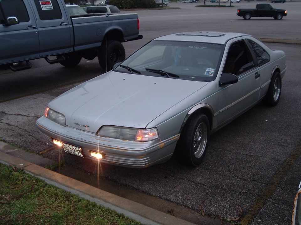 Silver Ford Thunderbird coupe with lights on parked at a curb at dusk