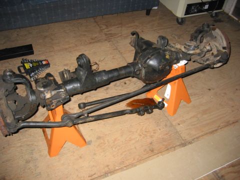 1991 Ford Explorer - Complete front axle assembly propped on two orange jack stands on a plywood shop floor, with the differential housing centered and knuckle,…