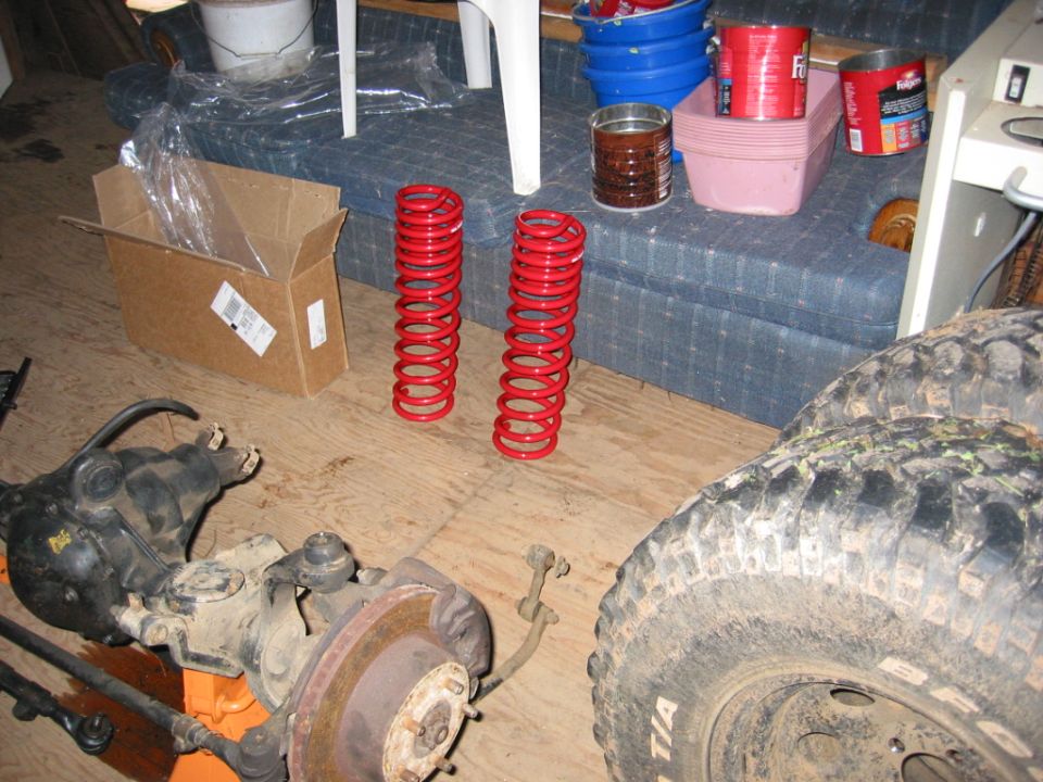 Two red coil springs, a front axle assembly, and a large tire on a wooden floor