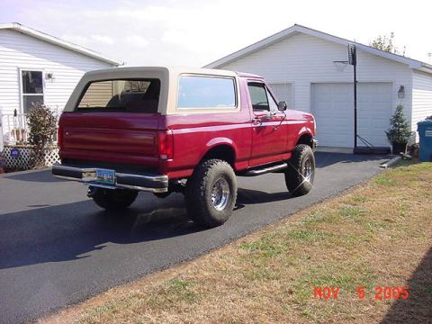 1989 Ford Bronco - Three-quarter rear view of a lifted red two-door Ford Bronco with a tan removable hardtop, parked in a paved driveway beside a white house…