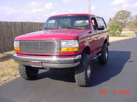 1989 Ford Bronco - Low front three-quarter view of a lifted red two-door Ford Bronco on a paved drive beside a wood privacy fence, sunlight glinting off its c…