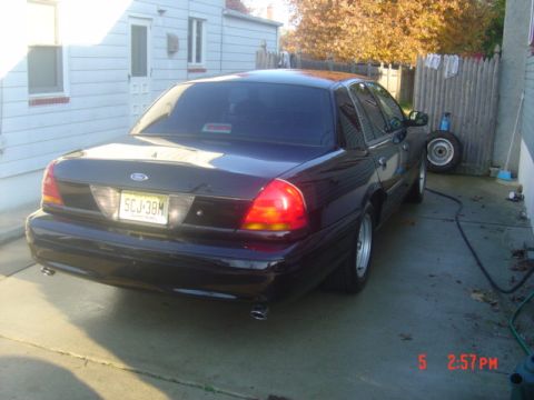 1999 Ford Crown Victoria - A dark Ford Crown Victoria is parked in a driveway next to a house and a fence.