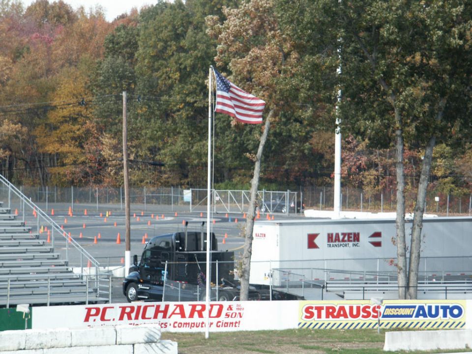 Black truck and white Hazen Transport trailer parked near bleachers with an American flag flying