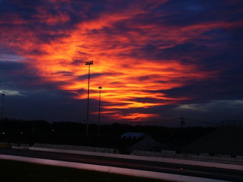 2001 Mercury Grand Marquis - Dramatic sunset paints the sky with vibrant orange and red hues over a darkened racetrack, illuminated by stadium lights.