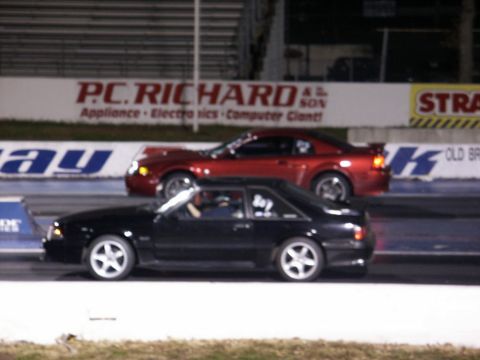 2001 Mercury Grand Marquis - Two cars compete on a drag strip, with a black 1990s Ford Mustang in the foreground and a red car behind it.