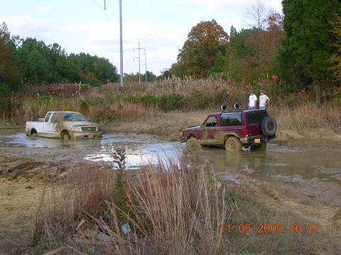 1989 Ford Bronco II - Two off-road vehicles are stuck in a muddy area with trees and tall grass in the background.