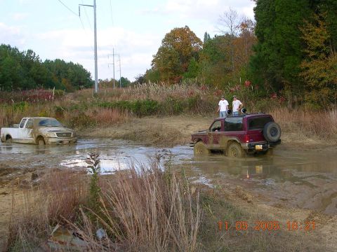 1989 Ford Bronco II - Two vehicles are stuck in a muddy area with trees and power lines in the background.