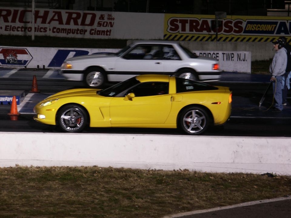 Yellow Chevrolet Corvette C6 coupe in motion on a drag strip, with a white Ford Mustang in the background