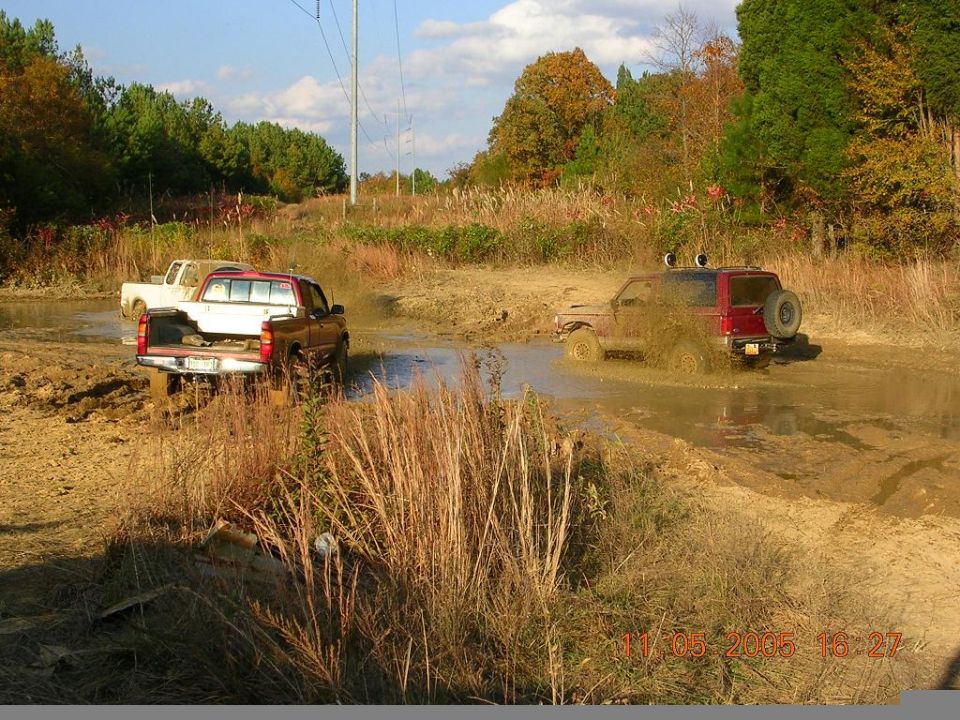 Trucks and an SUV driving through a muddy field with trees in the background
