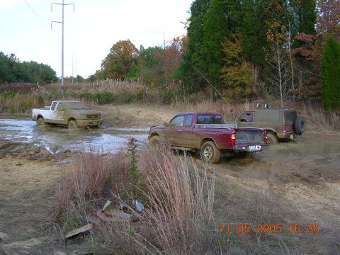 1989 Ford Bronco II - Multiple trucks navigate a muddy off-road course, with one white truck partially submerged in water and mud.