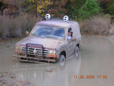 1989 Ford Bronco II - A person in a 1989 Ford Bronco II is stuck in a muddy body of water, with trees in the background.