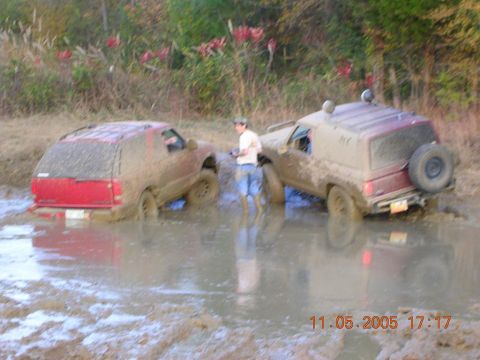 1989 Ford Bronco II - Two off-road vehicles are deeply stuck in mud and water, with a person standing between them.