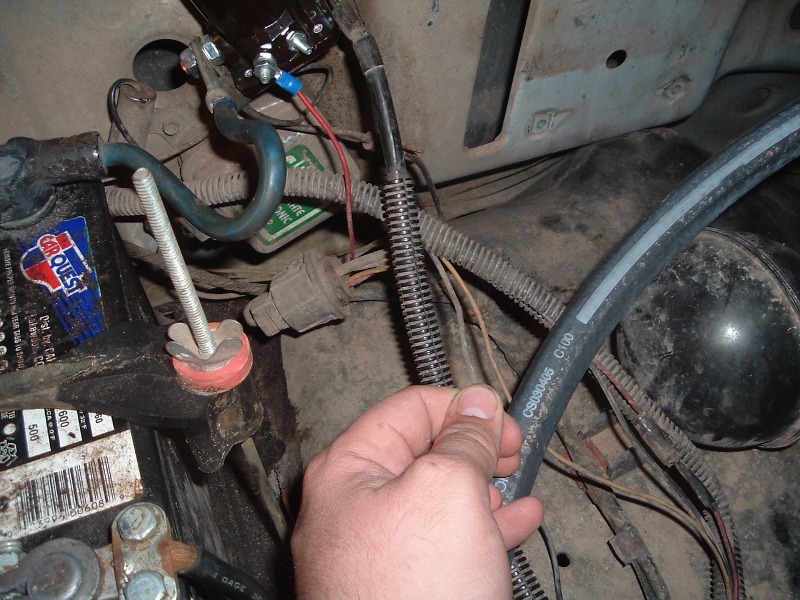 Close-up of a vehicle's engine bay showing the battery, wiring, and a hand holding a hose