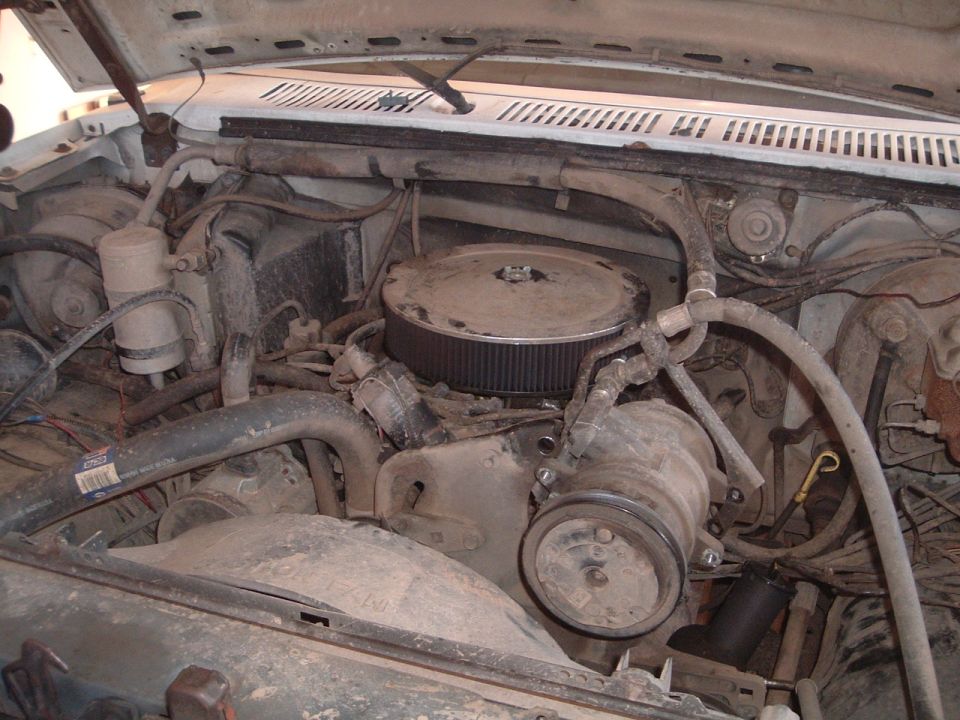 Engine bay of a white 1980s Ford truck with a V8 engine, air filter, and hoses. The engine is dusty and appears to be in a used condition