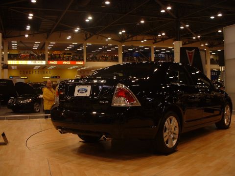1999 Ford Crown Victoria - A black Ford sedan is displayed at an auto show, with concessions signage in the background.