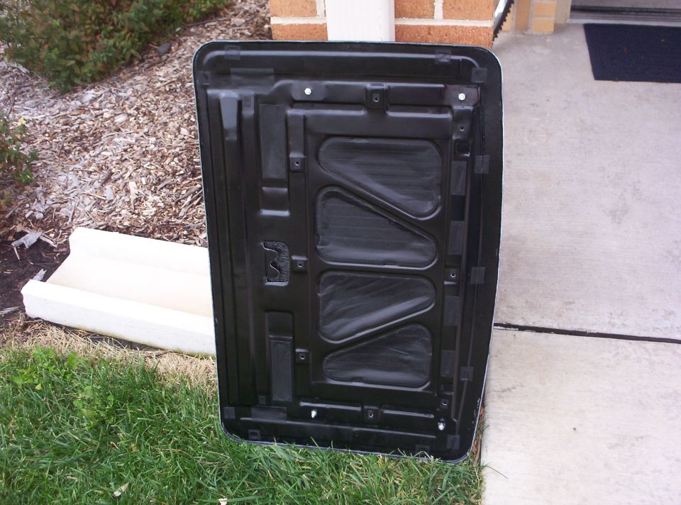 Underside of a black car hood, showing structural ribs and insulation, outdoors on grass and concrete