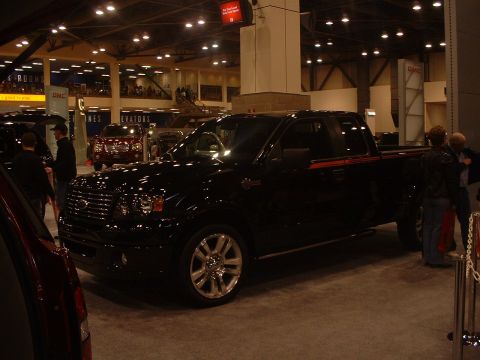 1999 Ford Crown Victoria - Black pickup truck on display at an auto show, with other vehicles and people in the background.