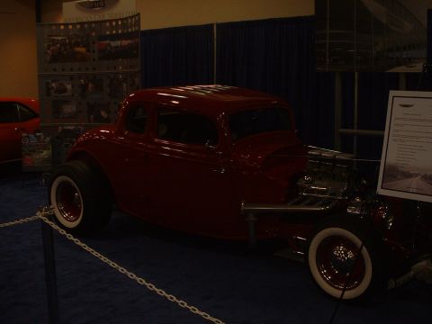 1999 Ford Crown Victoria - A red hot rod coupe is displayed indoors with a sign next to it and another car partially visible on the left.