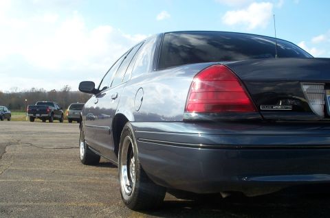 1998 Ford Crown Victoria - A dark blue sedan is parked on an asphalt lot, with other vehicles visible in the distance.