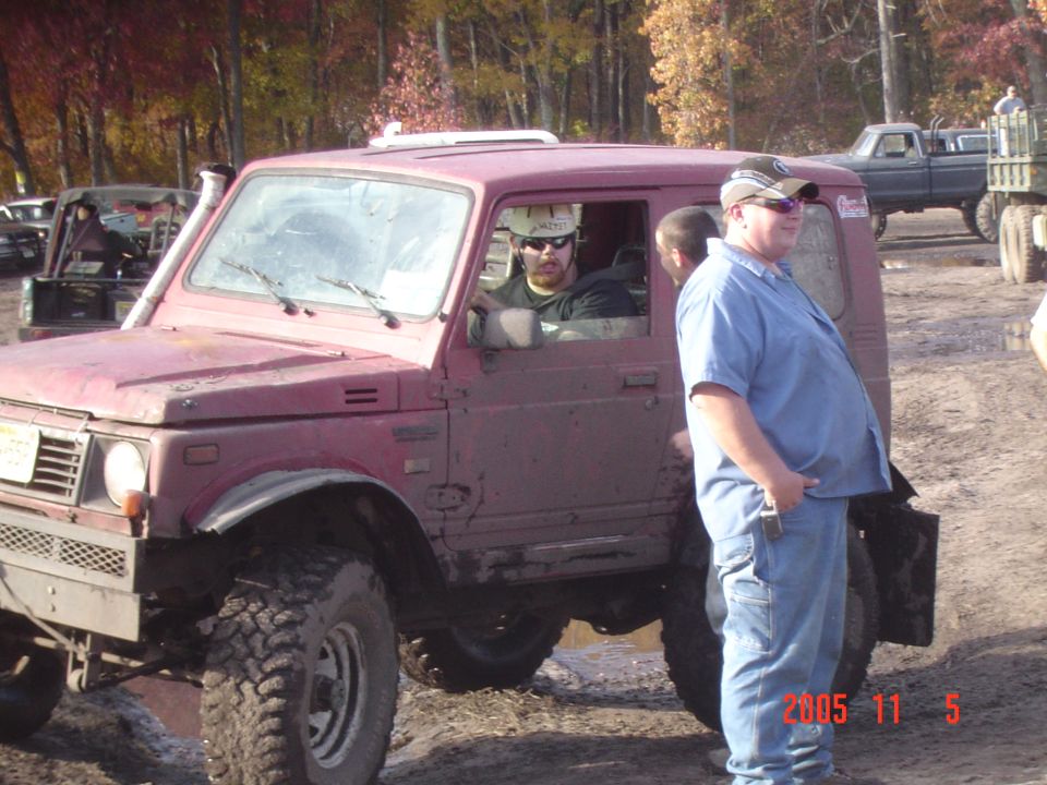 Maroon Suzuki Samurai with people inside, parked in mud. Another truck is visible in the background