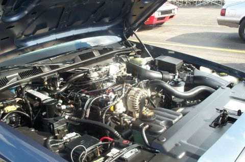 1998 Ford Crown Victoria - The engine compartment of a 1998 Ford Crown Victoria is displayed, with other vehicles parked nearby.