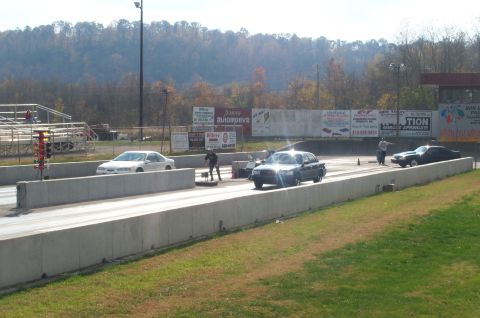 1998 Ford Crown Victoria - Two cars race down the drag strip on a sunny day, with trees and grandstands visible.