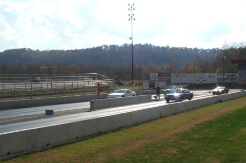 1998 Ford Crown Victoria - Cars line up on a drag strip under a bright sky with trees in the distance.