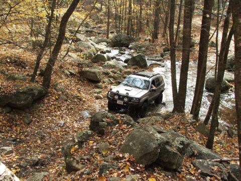 1996 Ford Bronco - A silver SUV drives through a shallow, rocky stream in a wooded area during autumn.