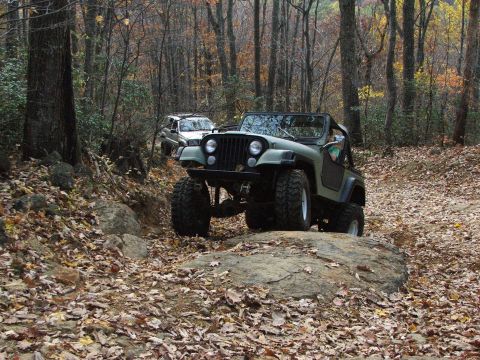 1996 Ford Bronco - An olive green Jeep Wrangler navigates a rocky trail in a wooded area, with another vehicle following behind.
