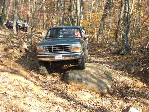 1996 Ford Bronco - 1996 Ford Bronco tackles a boulder on a forest trail with other vehicles present.