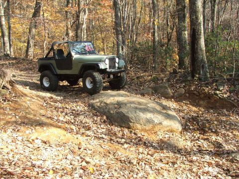 1996 Ford Bronco - A green Jeep Wrangler navigates a challenging off-road trail during the fall season, surrounded by trees with autumn foliage.