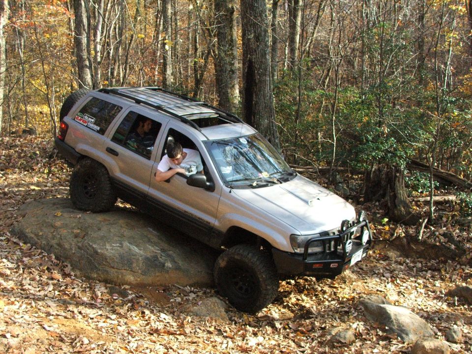 Silver Jeep Grand Cherokee with off-road tires climbing a rock in a wooded area